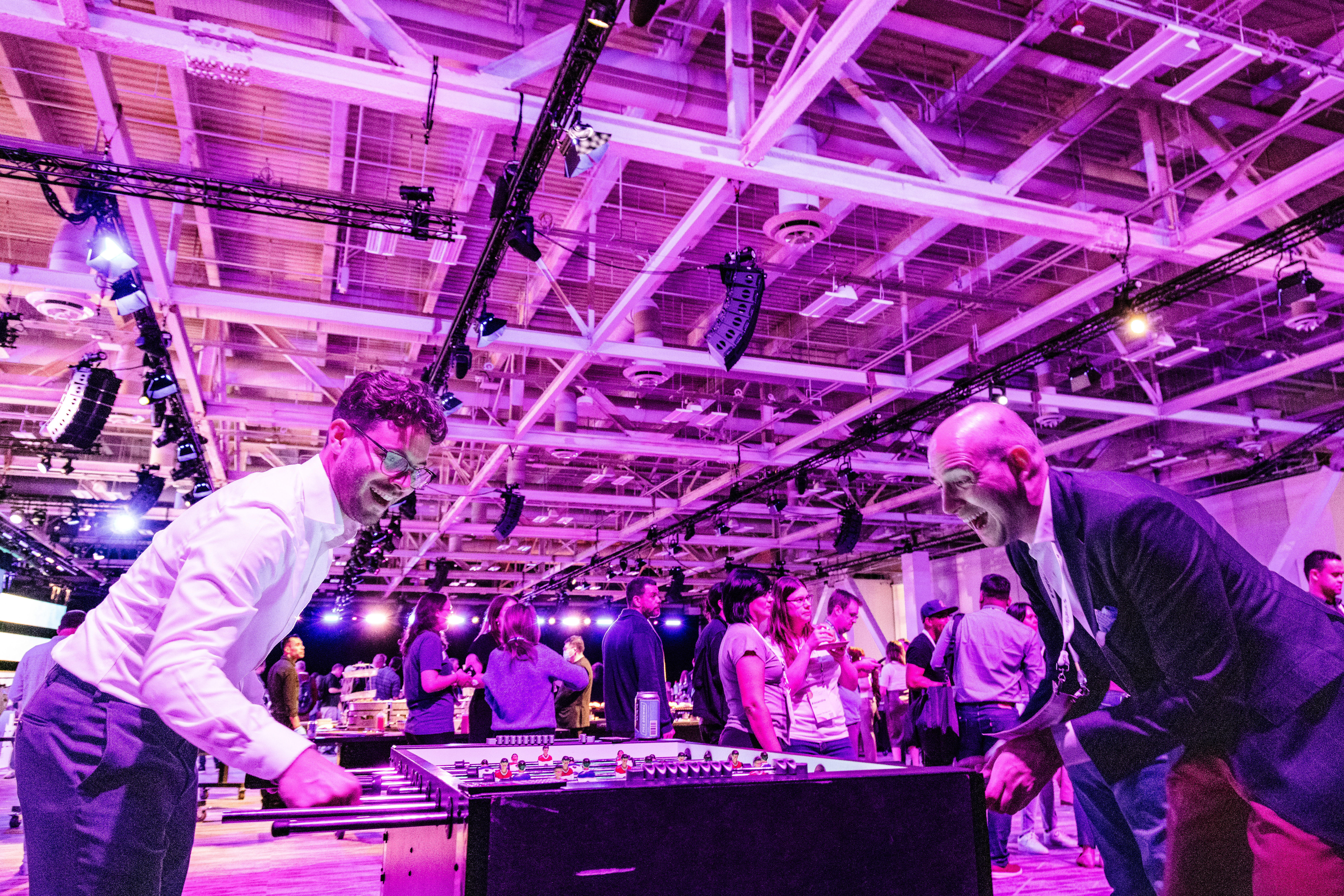 At the attendee party, two Smartsheet ENGAGE participants enjoy a game of table hockey while others mingle in the background.